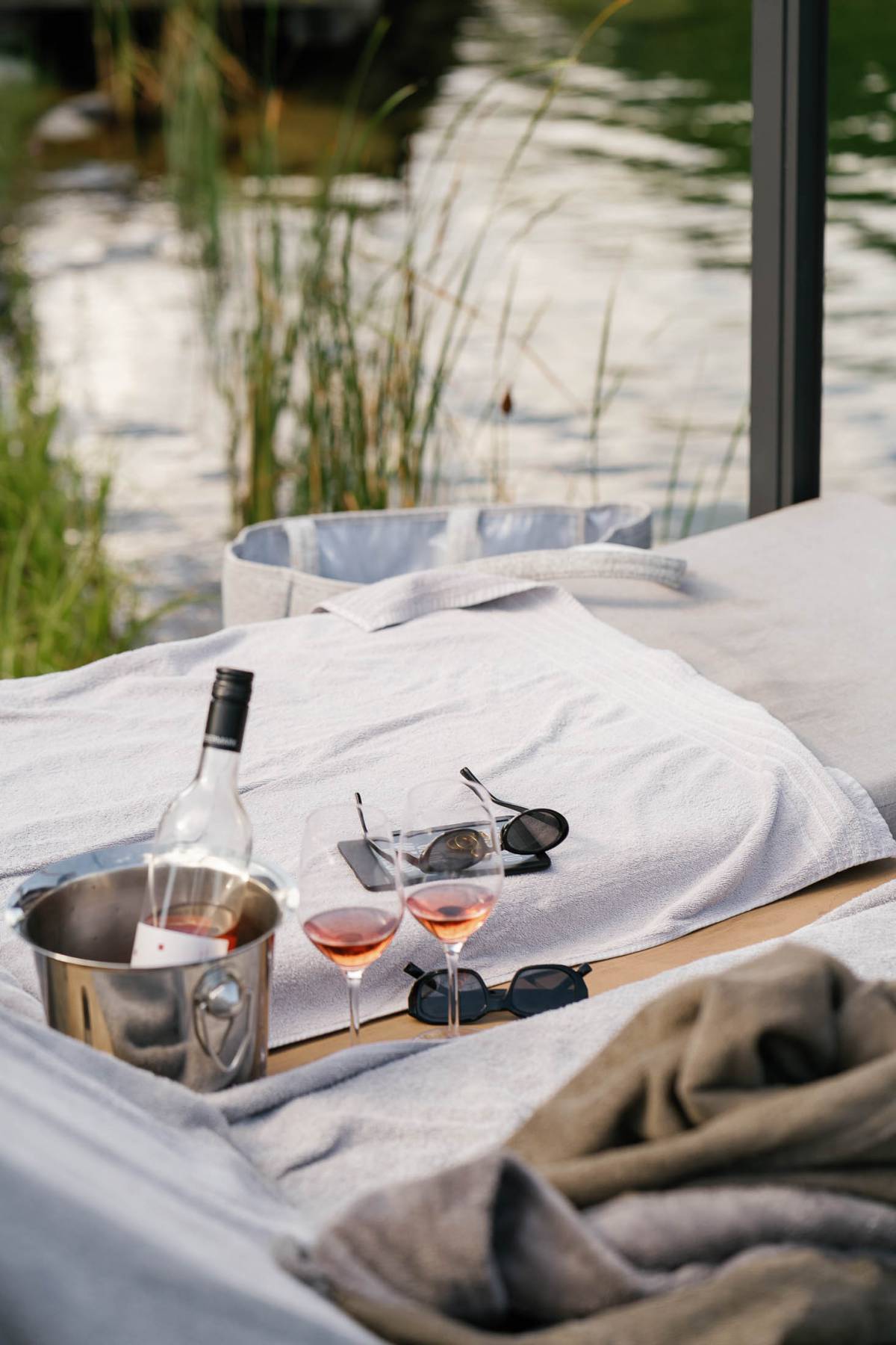 Nahaufnahme einer Sonnenliege am Wasser mit zwei Gläsern Roséwein, einer Weinflasche im Eiskübel, Sonnenbrillen und einem Smartphone