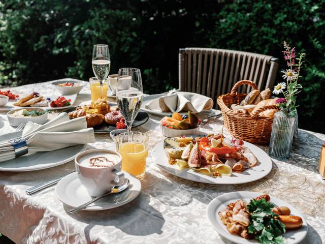 Een rijk gedekte ontbijttafel in de buitenlucht met koffiespecialiteiten, sappen, mousserende wijn, een schaal met vleeswaren en een mand vol broodjes