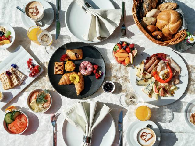Bovenaanzicht van een uitgebreide ontbijttafel met wafels, croissants, vleeswaren, vers fruit, sappen en koffie met melkschuimkunst in de vorm van een hart