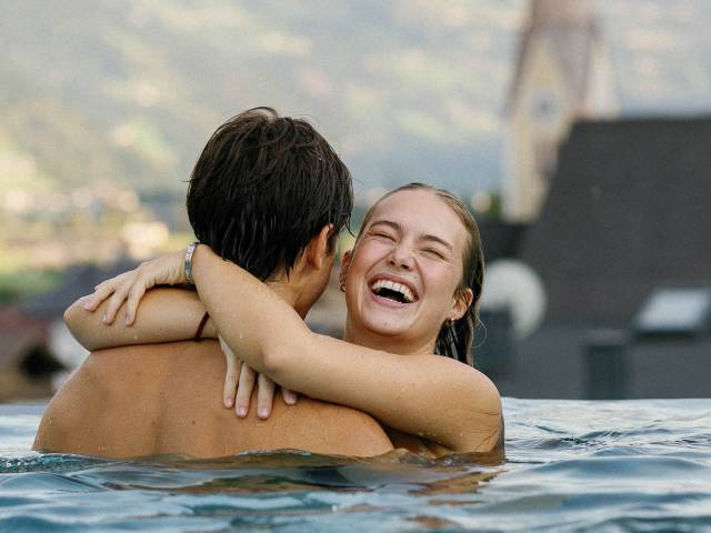 Een vrouw omhelst een man in een buiten-infinity pool en lacht hartelijk. Op de wazige achtergrond zijn bergen en een kerktoren te zien