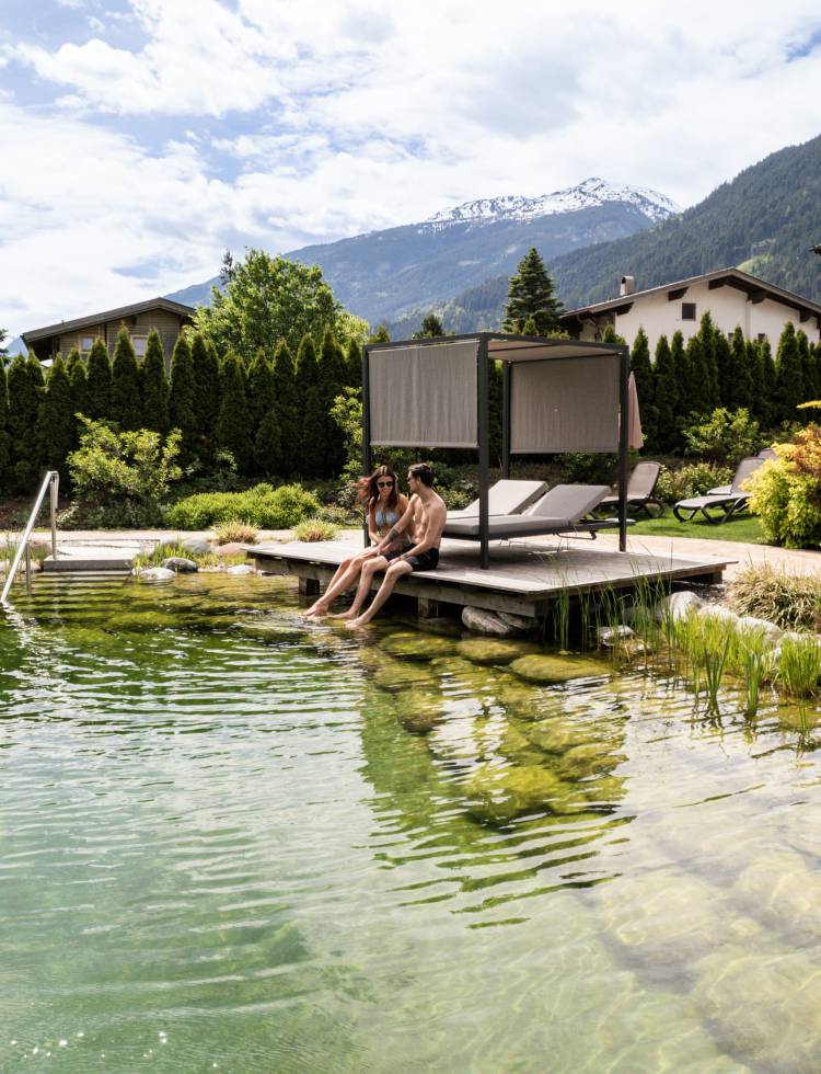 Natürlich gestalteter Badeteich mit Liegen und Pavillon, umgeben von Alpenpanorama