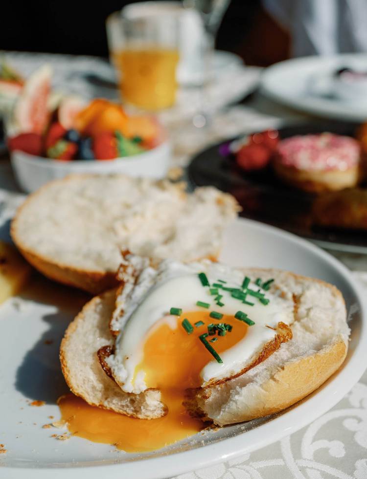 Close-up of a fried egg on a sliced bread roll with running egg yolk. In the blurred background, there are fruits and juice