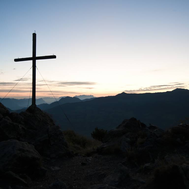 Silhouette eines Gipfelkreuzes vor den Bergen im Licht des Sonnenaufgangs