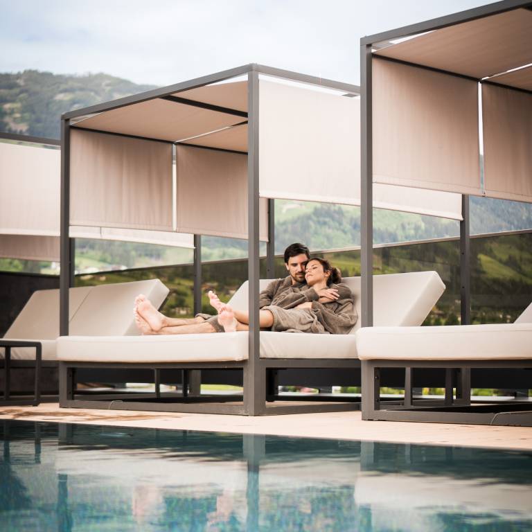 Entspanntes Paar kuschelt auf Liege am Pool mit Blick auf grüne Alpenlandschaft