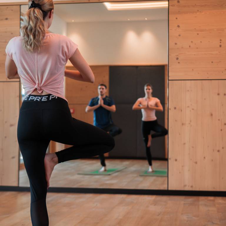 Gäste machen Yoga vor einem großen Spiegel im hellen Fitnessraum mit Holzwand und Yogamatte